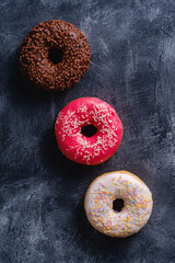 Chocolate, pink and vanilla donuts with sprinkles in row, sweet glazed dessert food on dark concrete textured background, top view