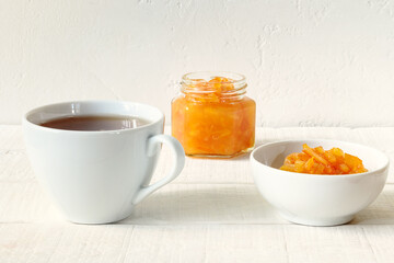 Cup of tee, glass jar and porcelain bowl with orange jam on a white wooden table