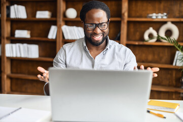 Confident hispanic man with handsome stubble, talking in front of computer, wearing blue shirt,...