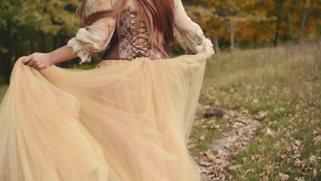 Happy joyful beautiful young woman in a medieval dress is runing in forest, enjoying nature. Slow motion. brown beige vintage dress. Long red hair fly flutter in wind. Cheerful girl back rear view.