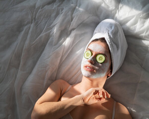 A woman with a towel on her hair and in a clay face mask and cucumbers in front of her eyes lies on...
