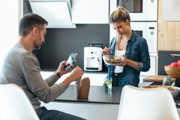Serious and unhappy young couple eating while using smartphone in the kitchen at home.