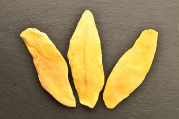 Three light yellow dry mango slices on a slate board, close-up, top view.