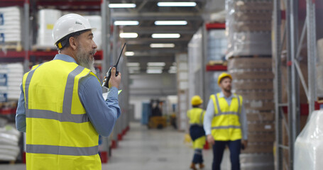 Senior manager in safety uniform transferring message to employee with walkie-talkie in warehouse