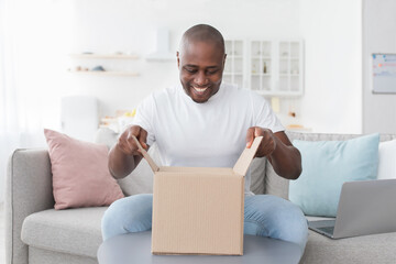 Happy black man unpacking delivery box, sitting on couch at home interior. Satisfied customer...