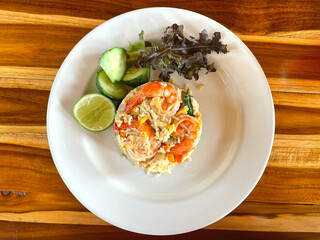 Shrimp fried rice topped with Placed on a white plate with vegetable, cucumbers and lemon beside the plate With a wooden floor as the background. Top view of shrimp fried rice