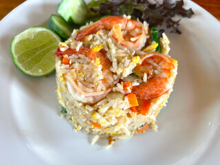 Shrimp fried rice topped with Placed on a white plate with vegetable, cucumbers and lemon beside the plate With a wooden floor as the background. Top view of shrimp fried rice