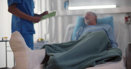 Doctor talking to senior man patient with broken leg lying in hospital bed