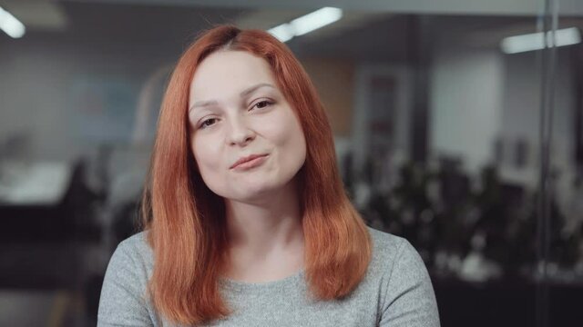Red-haired woman looking at camera and talking, blurred office on background. Online business meeting or job interview with participants using webcams. Concept of distance communication