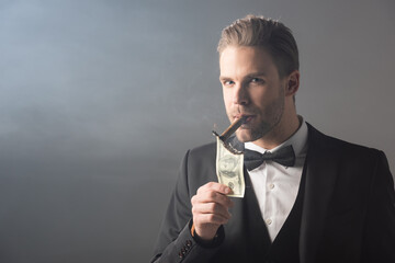 businessman lighting cigar from dollar banknote while looking at camera on grey background with smoke