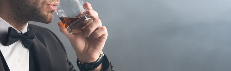 cropped view of businessman in bow tie drinking whiskey on grey background with smoke, banner
