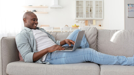 African american man reclining on couch in living room, using newest laptop, checking social media, panorama