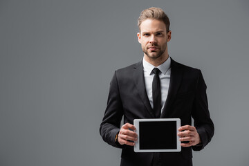 serious businessman in black suit showing digital tablet with blank screen isolated on grey