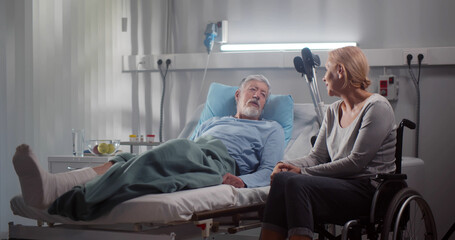 Senior woman in wheelchair visiting husband with broken leg in hospital ward