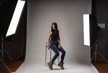 Alternative model with tattoos and jewelry poses sitting in a chair in a professional photo studio with spotlights and white background