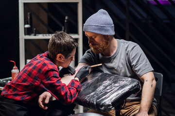 Girl tattoo artist makes a tattoo on the arm of a red-bearded hipster using the handpoke method in a tattoo parlor.