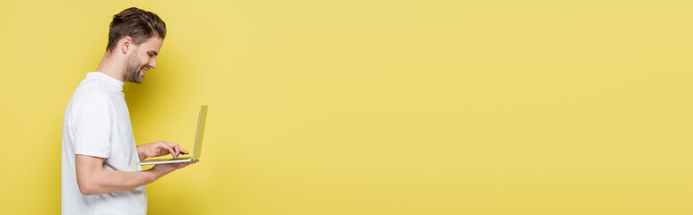 side view of smiling man typing on laptop on yellow, banner