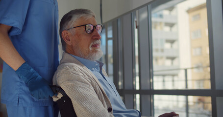 Senior man in wheelchair with nurse taking care looking through hospital window.
