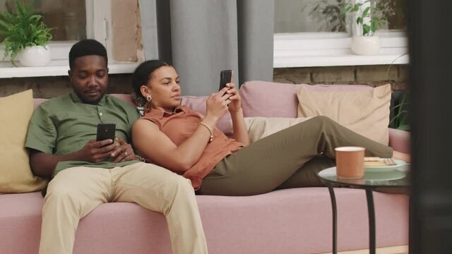 Medium shot of afro man and mixed race woman chilling on sofa together both using their smartphones scrolling feed and checking social networks