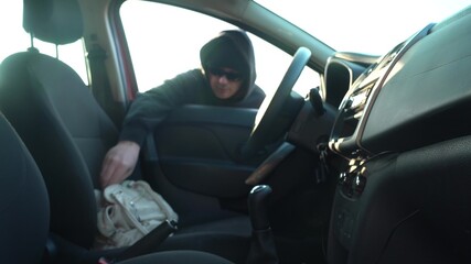a man in a black hoodie and black glasses steals a bag from the seat of someone else's car....