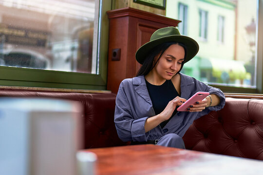 Stylish fashionable elegant cute attractive hipster woman traveler wearing felt hat using a phone resting and sitting by the window at cafe shop