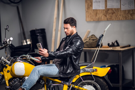 A motorcycle rider in a leather jacket with a smartphone in hand poses for social media and a video blog in a garage.