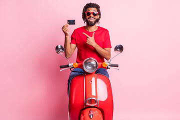 Portrait of handsome cheery funky guy riding moped demonstrating bank card advice isolated over pink pastel color background