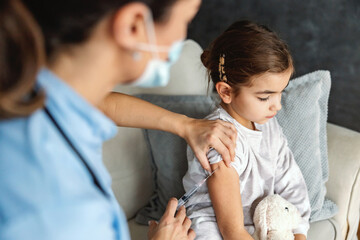 Obraz premium Ill little girl sitting on sofa at home and getting injection during covid outbreak. Doctors at home concept.