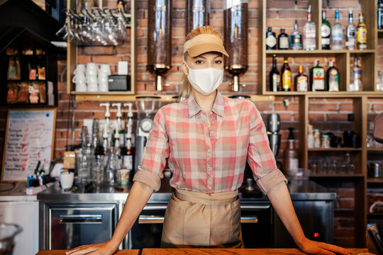 The Work Of A Bartender At The Time Of The Corona. Portrait Of A Female Person With A Mask Standing In A Bar And Wearing A Face Mask. She Expects To Order Coffee Or Cocktails During Covid 19