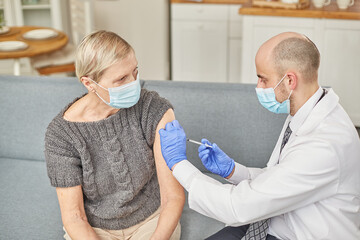 Man doctor vaccinates an elderly woman at her home wearing a protective mask. Vaccination concept