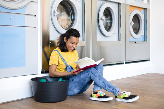 Latin woman reading a book sitting on the floor while waiting for clothes washing at the self-service laundry. She is smiling. - Powered by Adobe