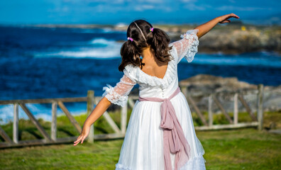 Fototapeta na wymiar Una niña de espaldas vestida de Primera Comunión juega con el viento con los brazos en cruz mirando hacia el Mar Cantábrico en el norte de España