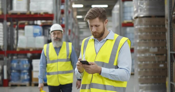 Aged warehouse manager giving helmet to young worker standing in using smartphone