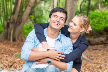 Romantic young couple hugging and laughing together while looking each other at outdoors. Cheerful woman blond hair embracing her boyfriend while sitting in the park. Valentines day concept.