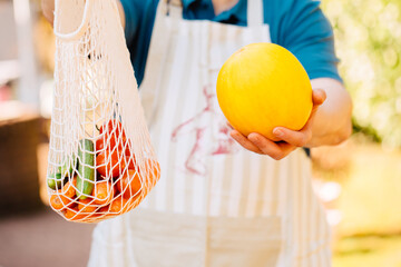 Weekly market vendor holds a honeydew melon and a reusable mesh bag with carrots, tomatoes and...