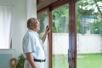 An elderly senior retirement man during isolation of a coronovirus living alone at home thinking and looking through window awaiting the end of a pandemic
