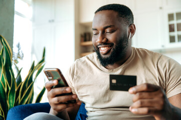 A satisfied smiling african american guy, in casual clothes, sits on the couch at home, holds a phone and a credit card in hands, pays for online purchases, enters a card number, online shopping