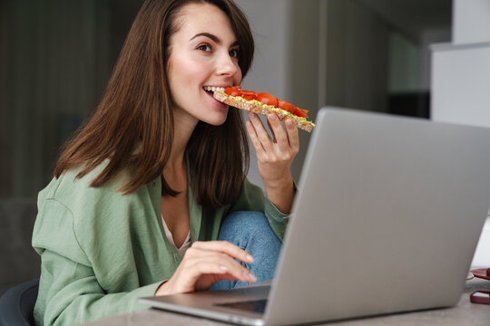 Young Happy Woman Eating Avocado Toast While Working With Laptop