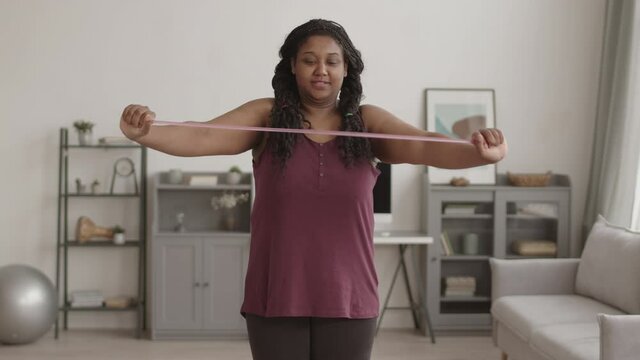 Lockdown of young curvy African-American sporty woman wearing sportswear standing in living room and doing strength exercise using elastic extender