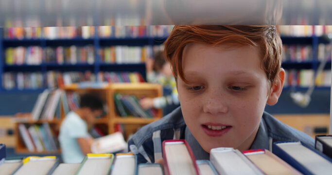 Close up of redhead cute schoolboy taking book from bookshelf in library