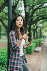 Naklejka premium charming young asian japanese woman standing by light pole in park pathway reading online map app information on mobile phone. cute girl holding cellphone point aside searching right way in garden.