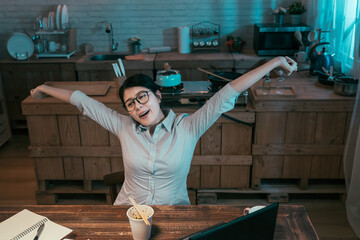 tired young office lady employee sitting at dark home kitchen in night and raising arms stretching...