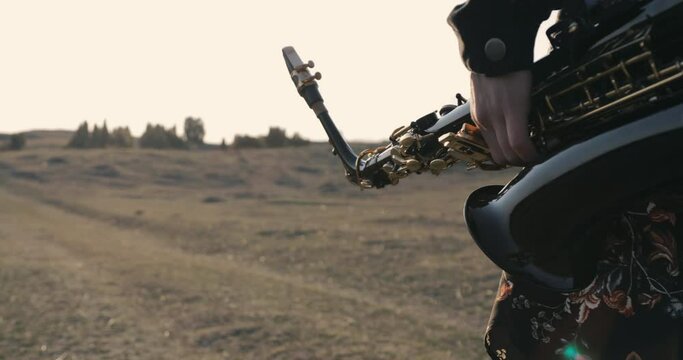 the girl carries a saxophone in her left hand across a meadow with low vegetation and the sun in the background