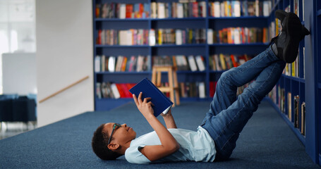 Afro-american boy lying on floor and reading book in modern school library