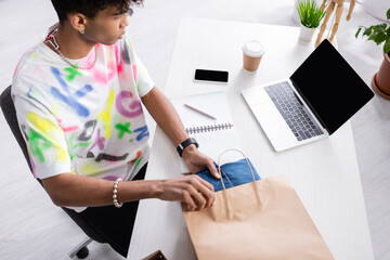 Laptop near african american owner of showroom putting clothes in shopping bag at table