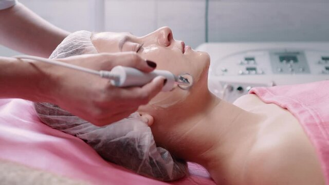 Skin care. Young woman at the reception at the beautician. The doctor is doing microcurrent therapy of the facial skin. Side view. Slider shot.