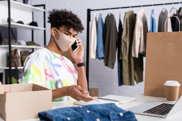 African american owner of showroom talking on smartphone and holding pen near clothes and packages...