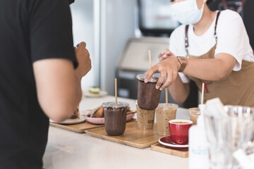 Waitress wearing protection face mask serving iced chocolate drink to customer in cafe.