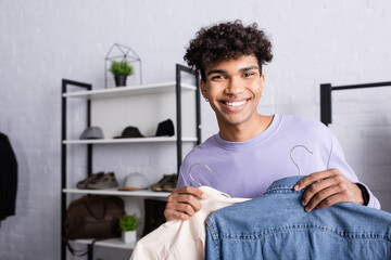 Arfican american businessman holding shirts on hangers and smiling at camera in showroom