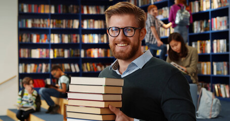 Bearded school teacher or librarian holding books in hands standing in library interior. - obrazy, fototapety, plakaty